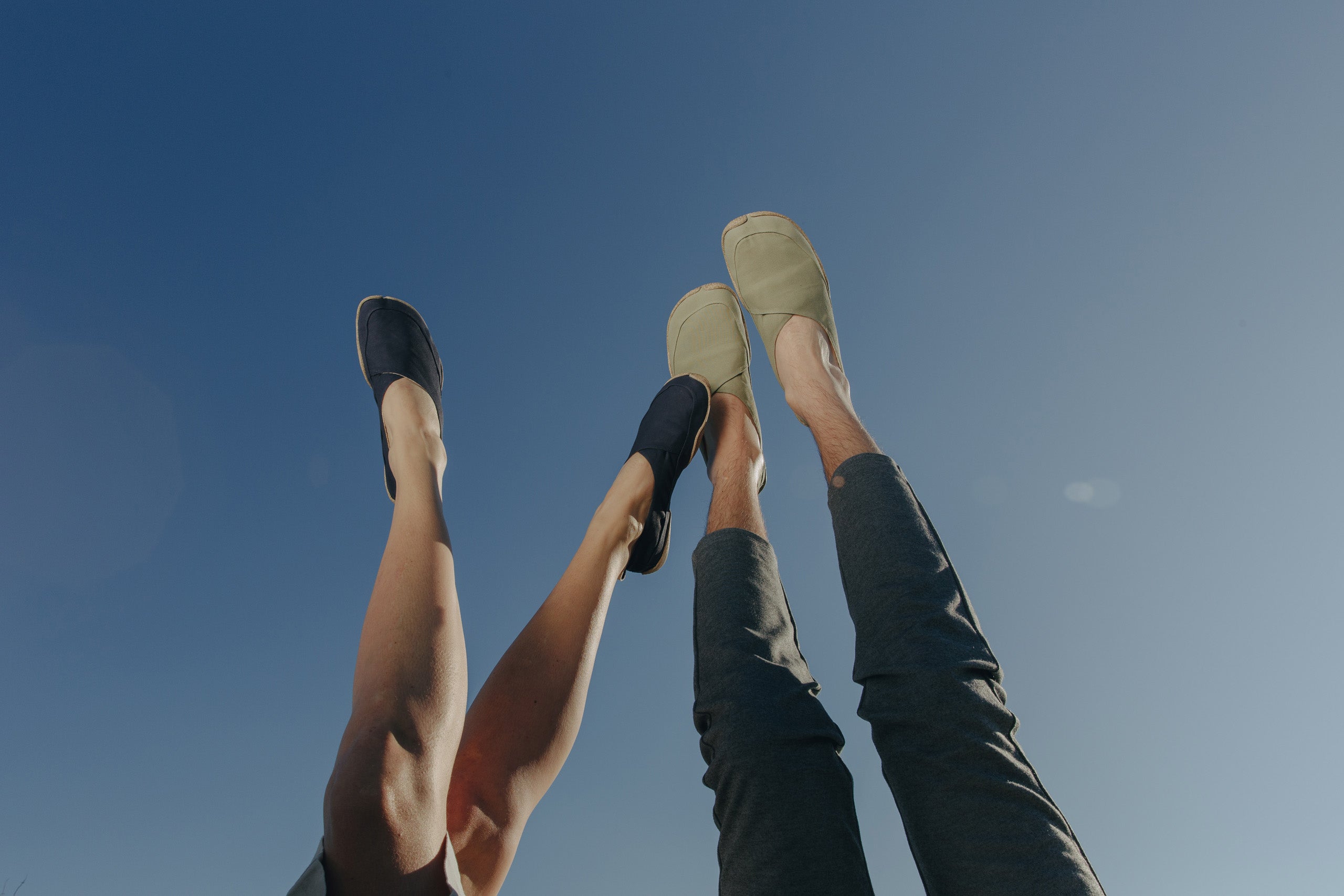 Two pairs of legs, up to the knees in the picture, stretched into the cloudless sky. On the feet Wildling Shoes minimal shoes.