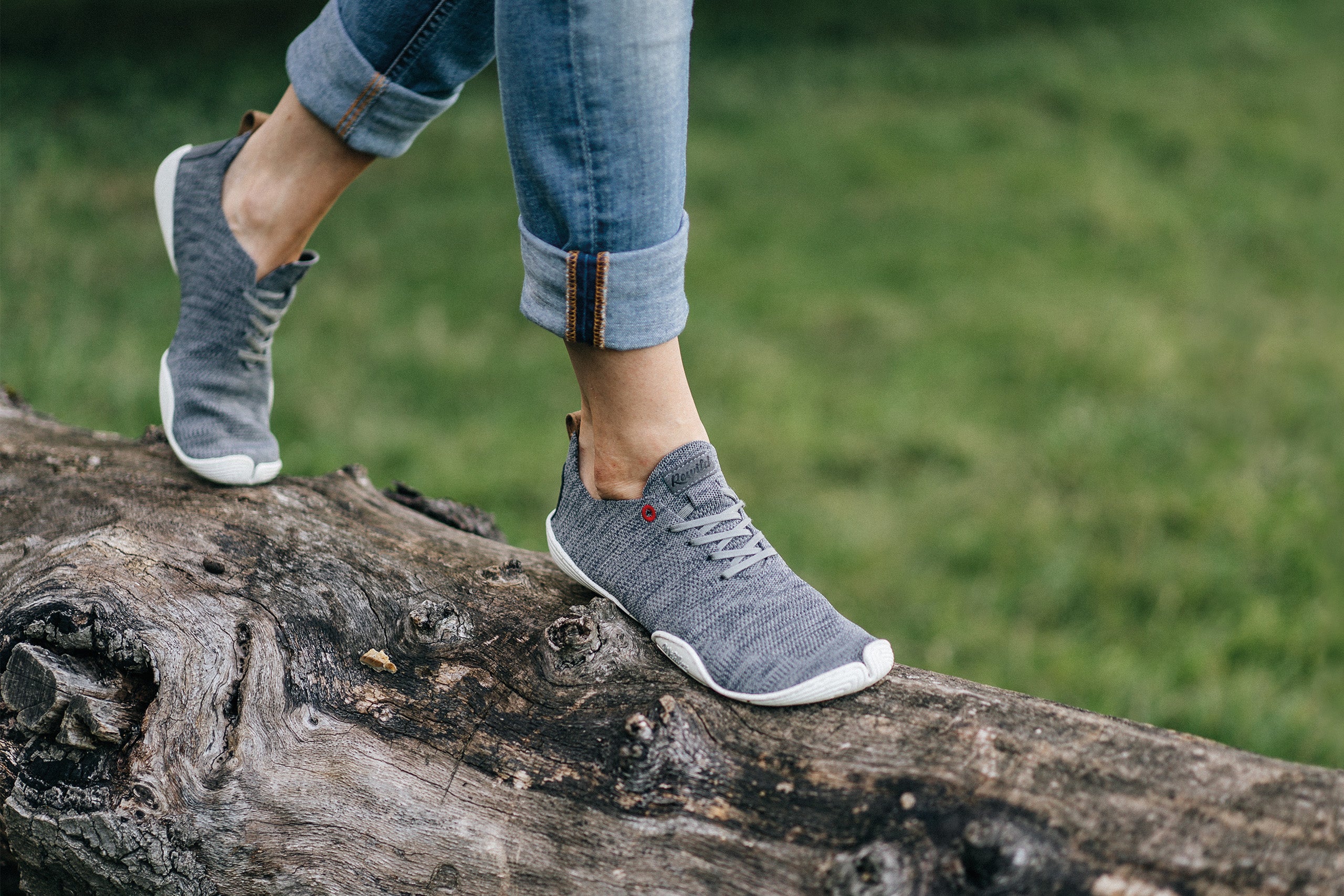 Two feet, somewhat up to the calves in the picture, balancing on a lying tree trunk; in the background a meadow; on the feet Wildling Shoes Minimal.