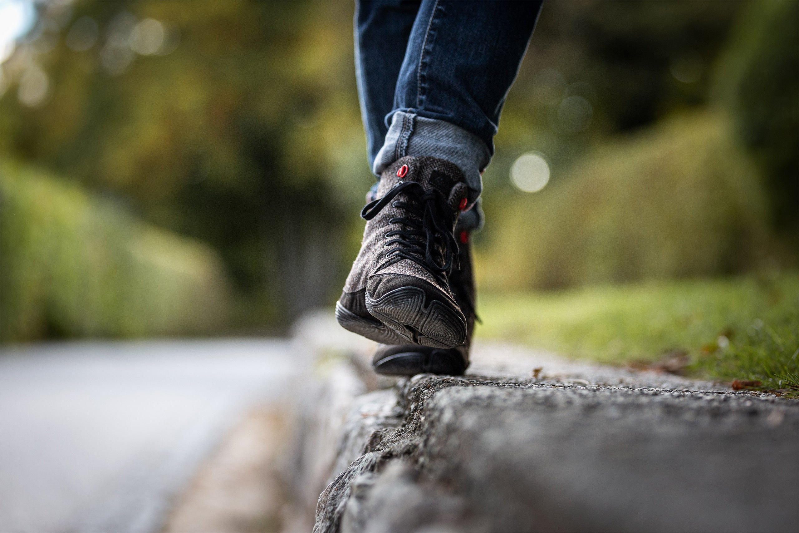 Two feet, slightly up to the calves in the picture, balancing on a lying tree trunk; in the background blurred a forest path and a piece of meadow; on the feet minimal shoes from Wildling Shoes.