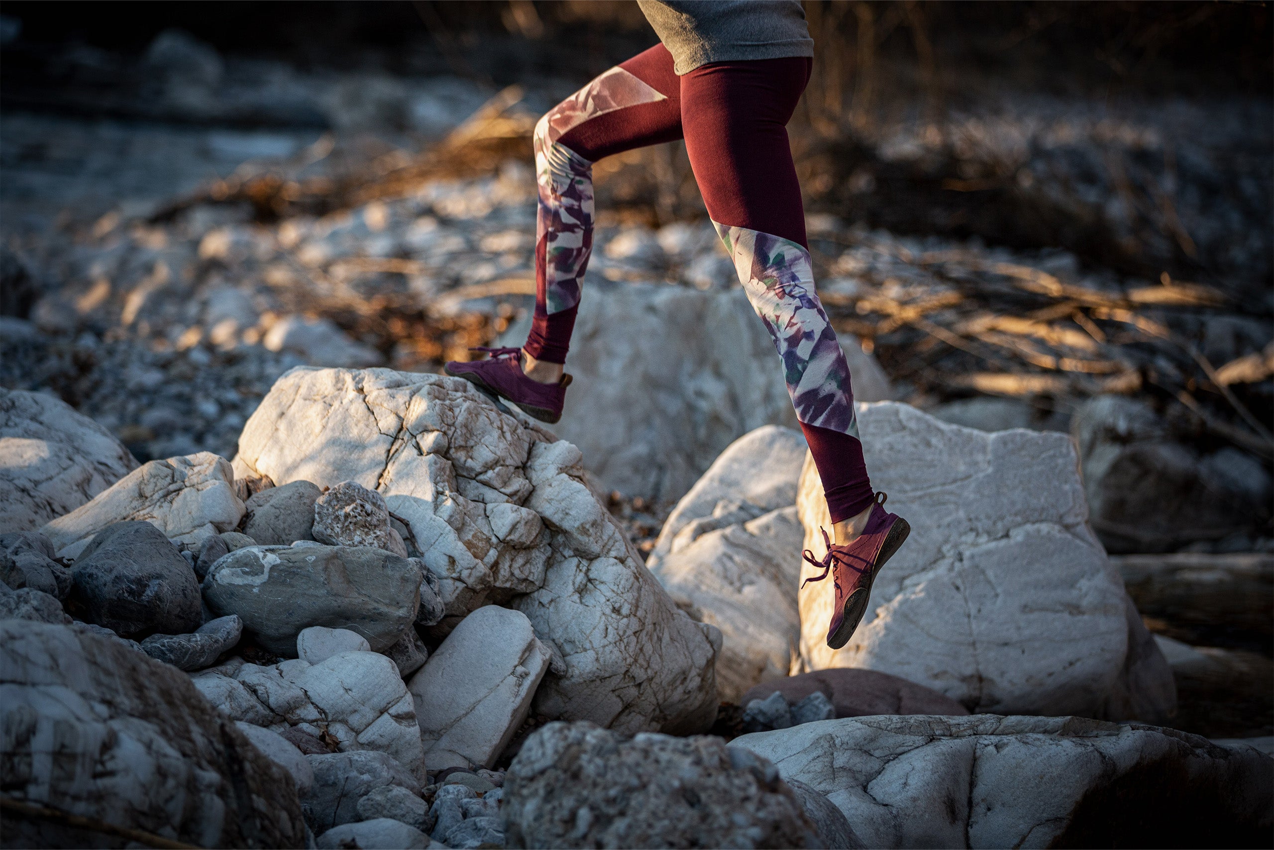 A person in dark red patterned leggings and Wildling Minimal shoes jumps over light colored boulders. The person is about up to the waist in the picture.