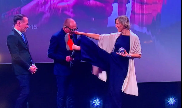 Die Übergabe des Deutschen Nachhaltigkeitspreises, Anna Yona auf der Bühne mit zwei Personen in schwarzen Anzügen, streckt ein Bein hoch bis auf Kinnhöhe, am Fuß ein leuchtend roter Wildling Shoes Minimalschuh. 